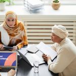 Três mulheres trabalham juntas em uma mesa com laptops, discutindo estratégias de publicidade digital em um escritório.
