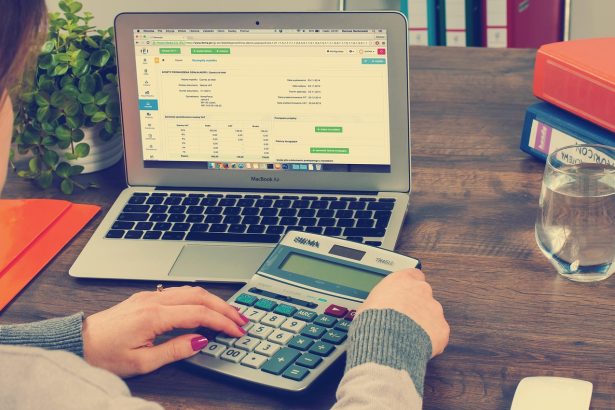 Mesa de escritório com computador portátil, pastas, documentos e calculadora, associados à contabilidade e à administração tributária