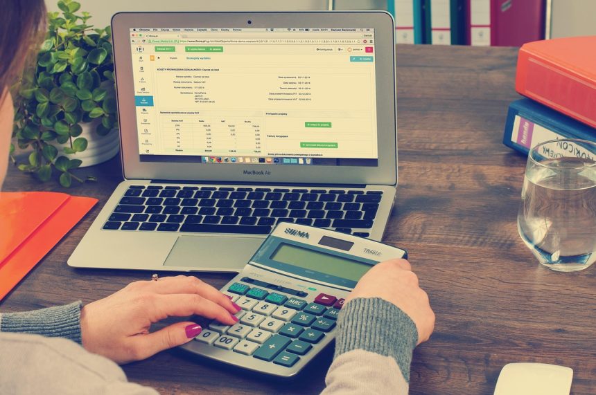 Mesa de escritório com computador portátil, pastas, documentos e calculadora, associados à contabilidade e à administração tributária