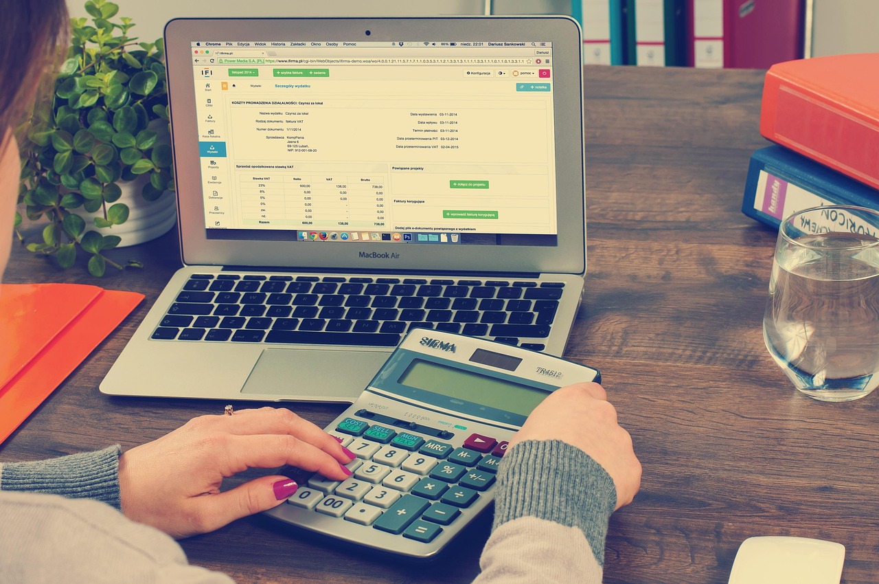 Mesa de escritório com computador portátil, pastas, documentos e calculadora, associados à contabilidade e à administração tributária